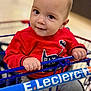 baby, child, shopping_cart, red_sweater, batman_logo, hand, face, smile, indoor, store, curious_eyes, grip, blue_handlebar, cute, young_child, casual_clothing, portrait, infant, closeup, happy