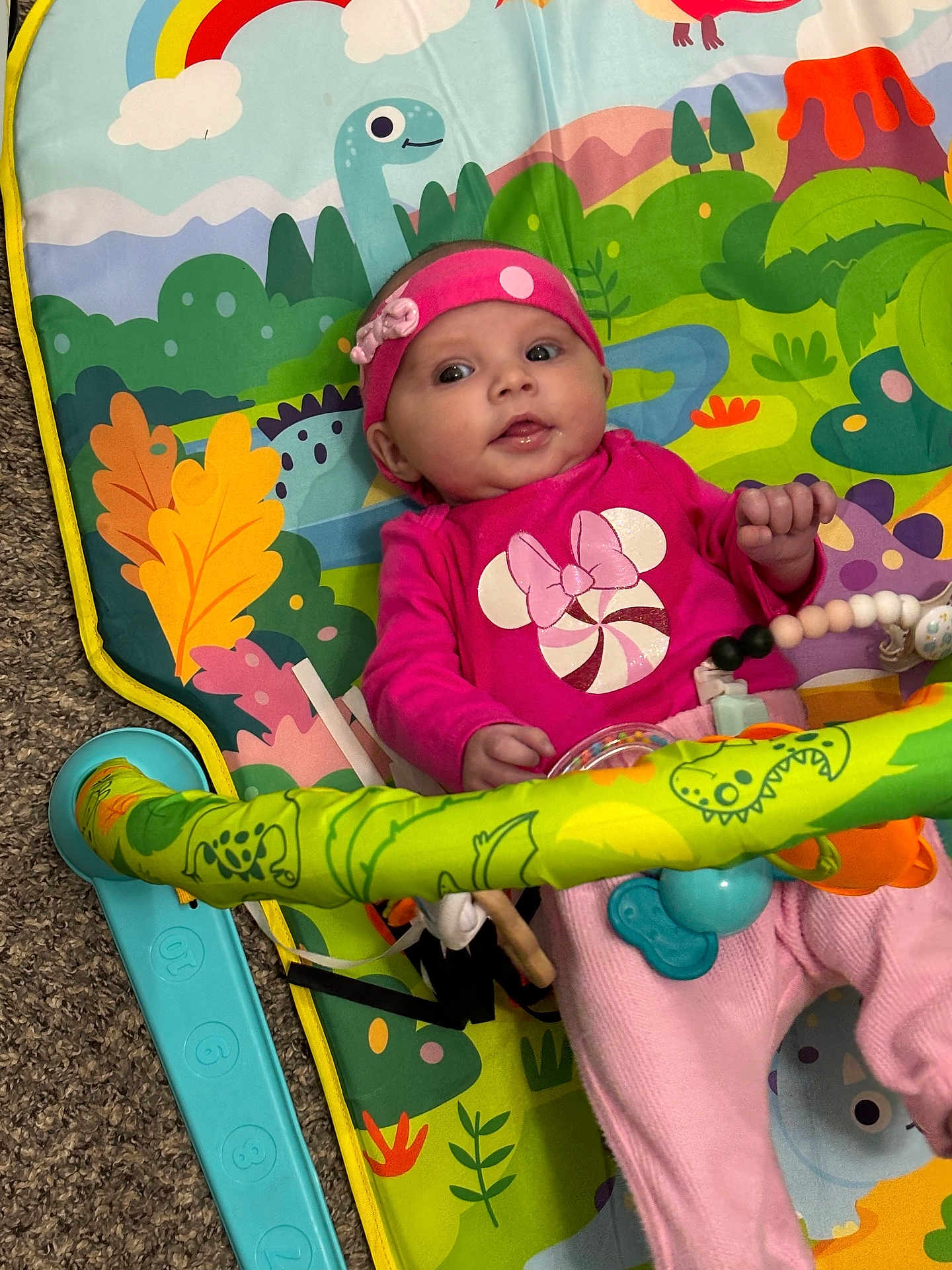 Tyler Sue is registered to the contest to win money with this photo: baby, carpet, child, colorful, cute, dinosaur_print, hand, headband, indoor, infant, legs, lying_down, pacifier_clip, pink_clothing, play_mat, portrait, smile, toy, toy_bar, wide_eyes