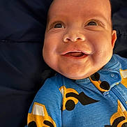 Kett is registered to the contest to win money with this photo: baby, infant, smile, face, onesie, clothing, blue, yellow, black, construction_vehicle_pattern, joy, expression, portrait, child, cute, happy, closeup, person, background, fabric