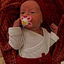 baby, infant, pacifier, red_clothing, white_clothing, blanket, newborn, face, headwear, lying_down, cute, small_hands, cozy, indoors, soft_texture, closeup, portrait, child, expression, resting