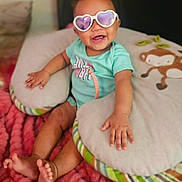 Carmella is registered to the contest to win money with this photo: baby, child, infant, smiling, sunglasses, heart_shaped_glasses, portrait, sitting, bed, pillow, blanket, plush_toy, monkey_toy, indoor, cozy, turquoise_shirt, pink_blanket, barefoot, happy, close_up