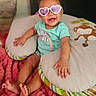 baby, child, infant, smiling, sunglasses, heart_shaped_glasses, portrait, sitting, bed, pillow, blanket, plush_toy, monkey_toy, indoor, cozy, turquoise_shirt, pink_blanket, barefoot, happy, close_up