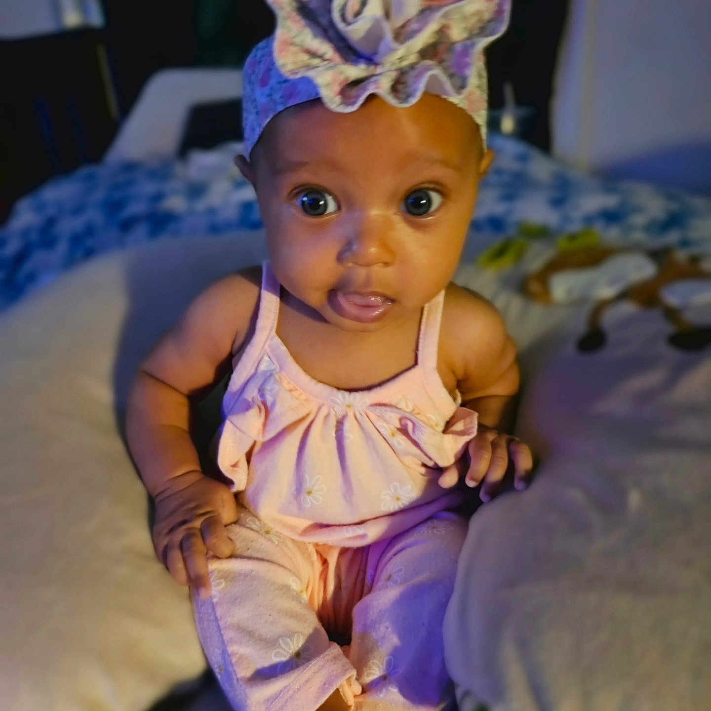Carmella is registered to the contest to win money with this photo: baby, infant, child, portrait, sitting, bed, bedroom, clothing, dress, hat, ruffled_hat, wide_eyed, looking_at_camera, indoor, home, blanket, bedding, colorful, cute, photography