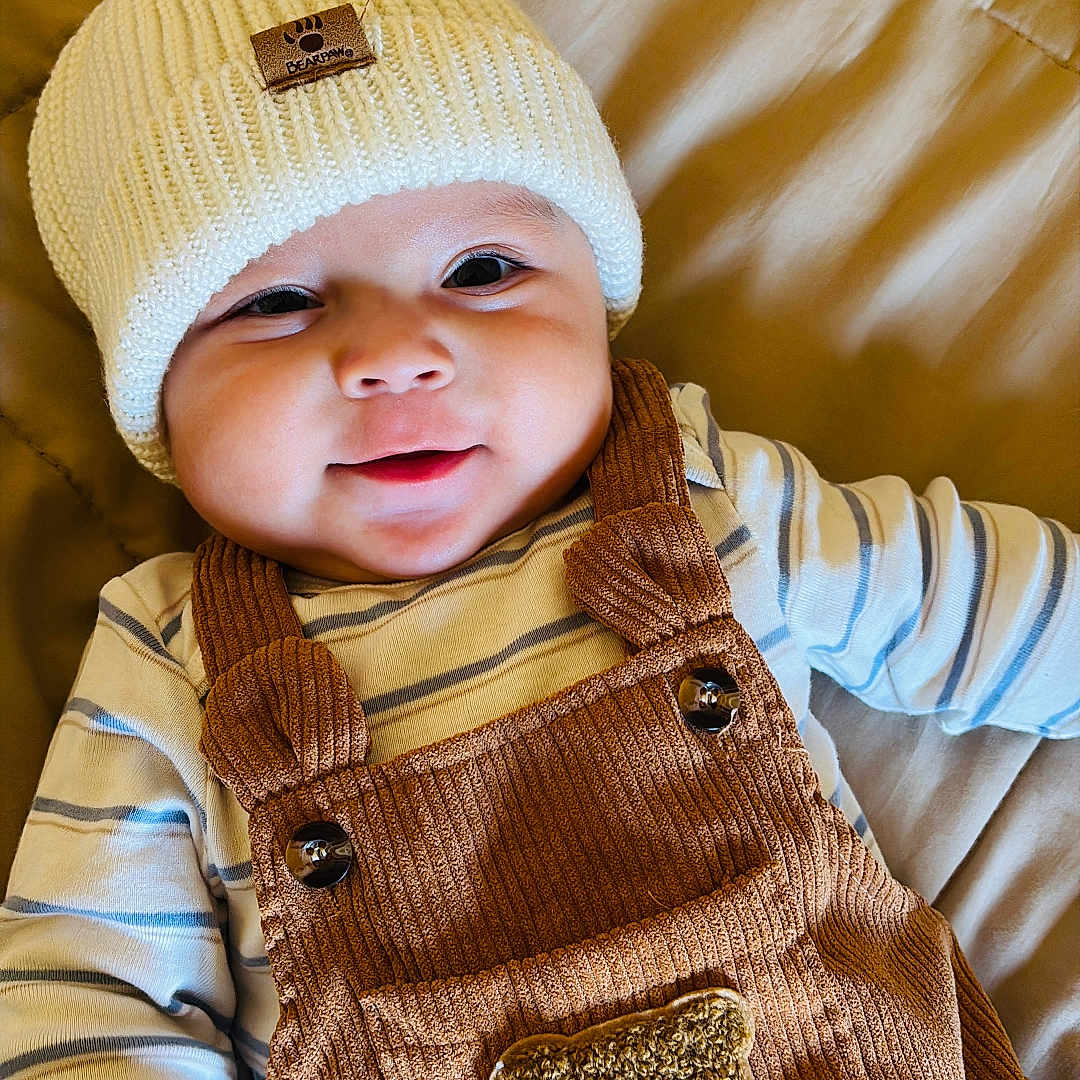 Johan is registered to the contest to win money with this photo: baby, beige_blanket, brown_overalls, child, cozy, cute, face, happy, hat, infant, knit_hat, lying_down, overalls, portrait, quilted_blanket, smiling, striped_clothing, striped_shirt, teddy_bear_patch, warm_clothing