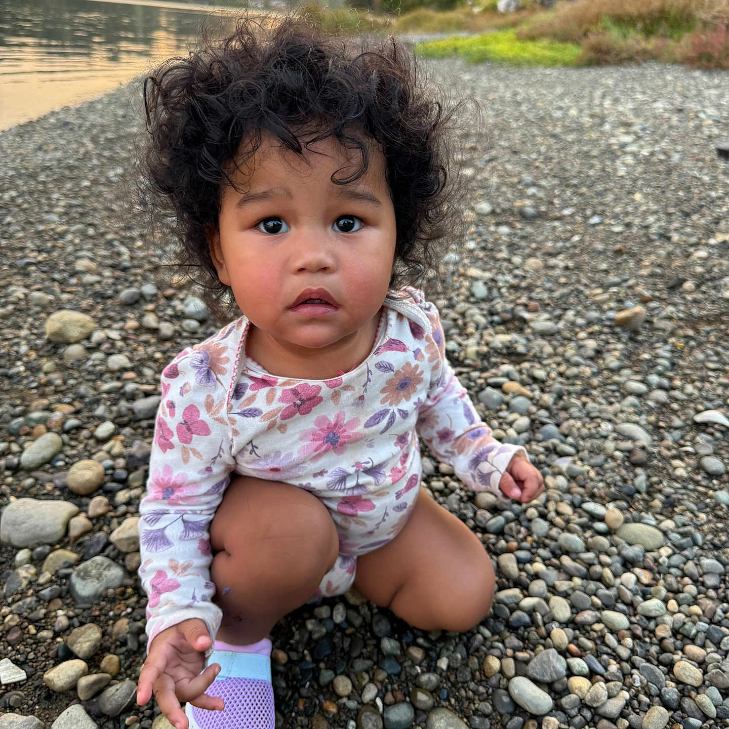 Aulani Rose joined the competition — help win amazing prizes! barefoot, child, crouching, curly_hair, expression, floral_clothing, grass, nature, outdoor, peaceful, person, portrait, riverbank, rocks, shoe, sunset, toddler, trees, water, young