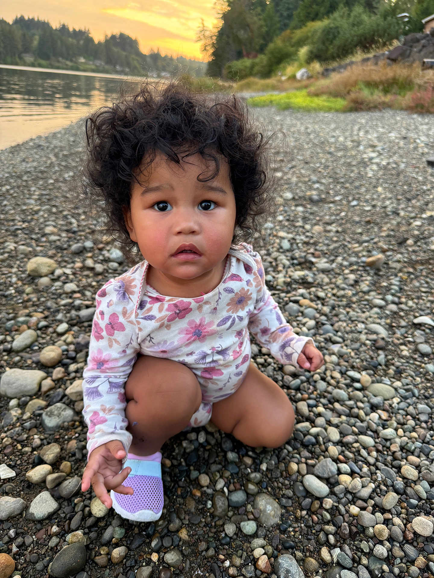 Aulani Rose joined the competition — help win amazing prizes! toddler, child, curly_hair, floral_clothing, riverbank, rocks, sunset, water, outdoor, nature, barefoot, shoe, expression, portrait, crouching, young, person, grass, trees, peaceful