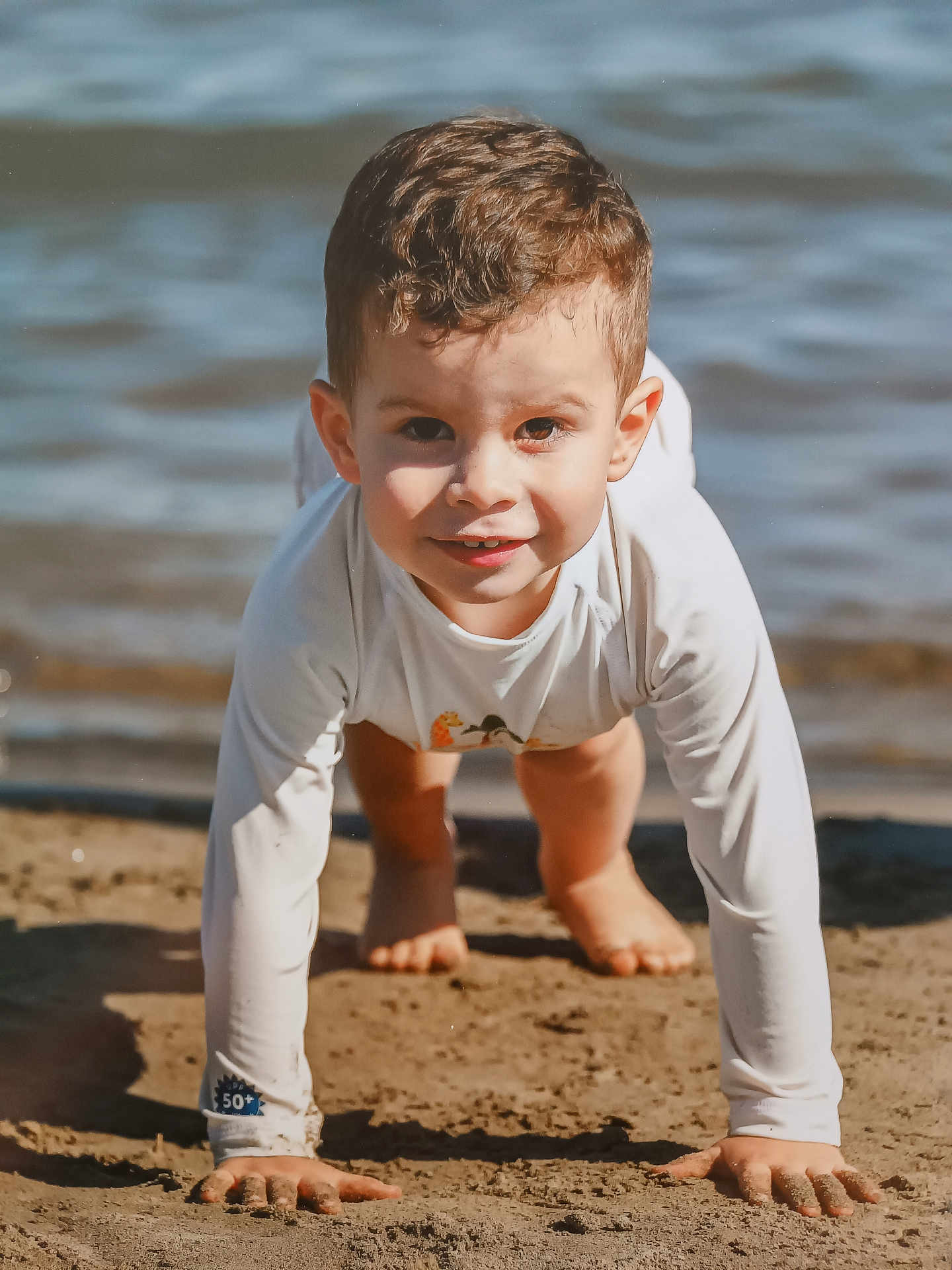 Diego a rejoint le concours — aidez-le/la à gagner de superbes lots ! child, sand, water, beach, crawling, smiling, sunlight, long_sleeve_shirt, barefoot, outdoor, playful, young_child, close_up, nature, daylight, happy, casual_clothing, curly_hair, face, skin