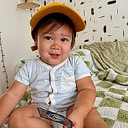 Desmond joined the competition — help win amazing prizes! toddler, child, yellow_cap, striped_onesie, card, indoor, blanket, green_and_white, cross_legged, smiling, face, home_decor, wall_pattern, dried_flowers, sitting, cute, playful, person, portrait, cozy