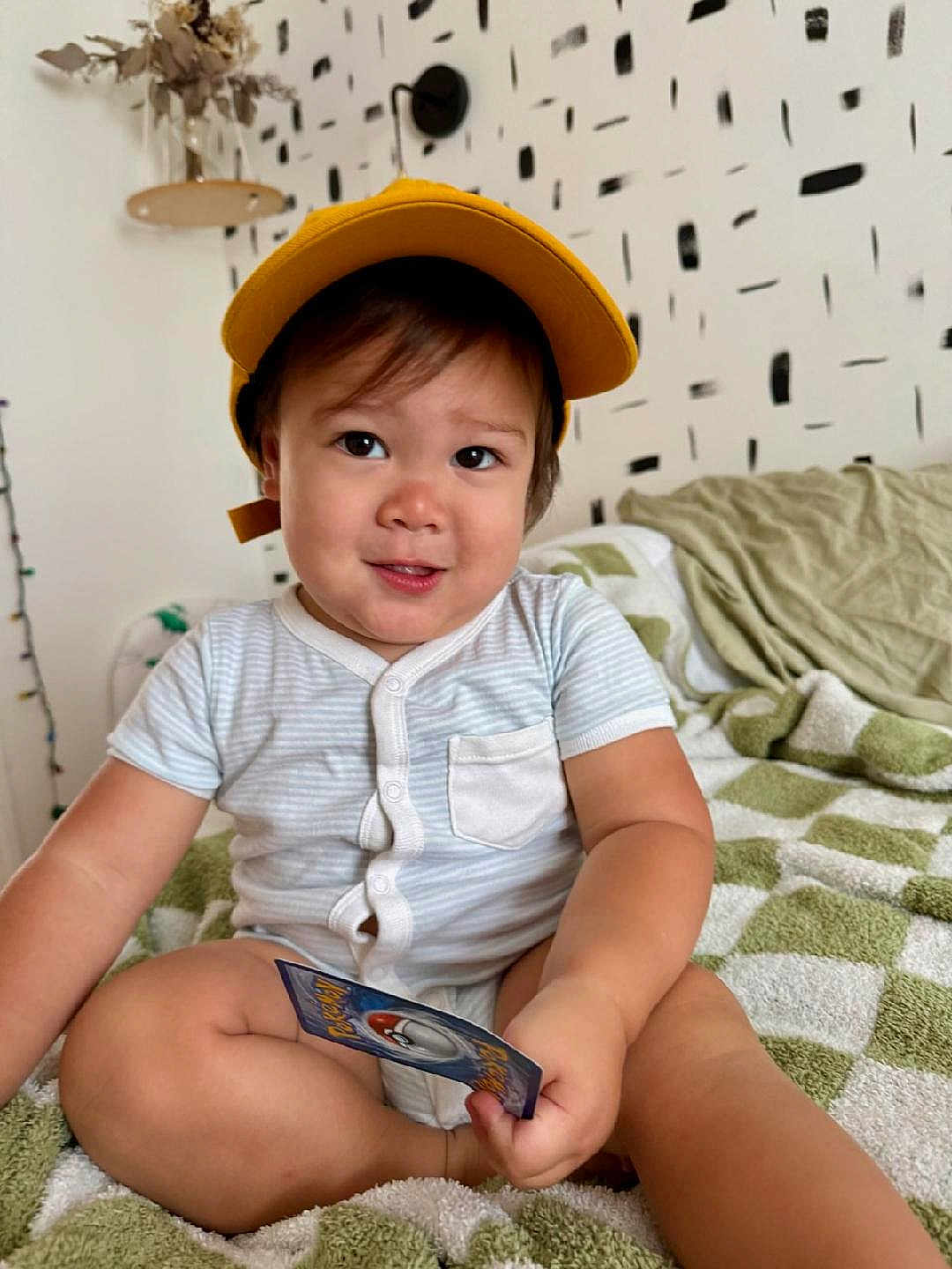 Desmond joined the competition — help win amazing prizes! toddler, child, yellow_cap, striped_onesie, card, indoor, blanket, green_and_white, cross_legged, smiling, face, home_decor, wall_pattern, dried_flowers, sitting, cute, playful, person, portrait, cozy