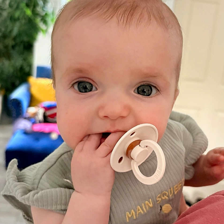 Brooke is registered to the contest to win money with this photo: baby, child, pacifier, person, indoor, couch, furniture, hair, ponytail, hand, face, smiling, clothing, room, window, plant, holding, cute, infant, portrait