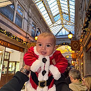 Leo is registered to the contest to win money with this photo: baby, child, santa_claus_costume, holiday, festive, lights, shopping_arcade, indoor, decorations, person, smiling, holding_hands, tongue_out, winter_clothing, celebration, christmas, face, happy, cute, festivity