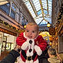 baby, child, santa_claus_costume, holiday, festive, lights, shopping_arcade, indoor, decorations, person, smiling, holding_hands, tongue_out, winter_clothing, celebration, christmas, face, happy, cute, festivity