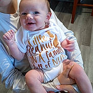 Amelia is registered to the contest to win money with this photo: baby, infant, smile, onesie, peanut, text, human_hand, person, indoor, floor, furniture, holding, happy, cute, child, young, skin, face, arm, legs