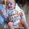baby, infant, smile, onesie, peanut, text, human_hand, person, indoor, floor, furniture, holding, happy, cute, child, young, skin, face, arm, legs