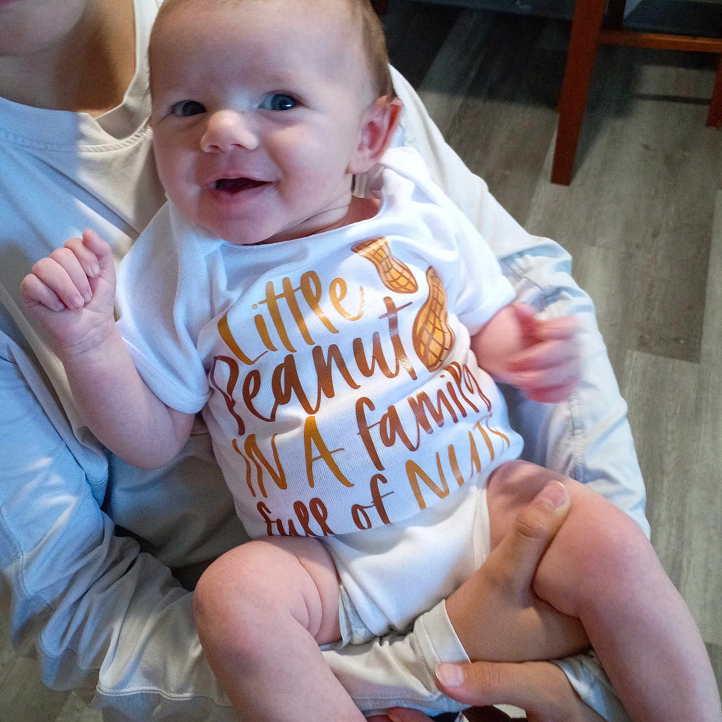 Amelia is registered to the contest to win money with this photo: baby, infant, smile, onesie, peanut, text, human_hand, person, indoor, floor, furniture, holding, happy, cute, child, young, skin, face, arm, legs