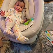 Gabriel is registered to the contest to win money with this photo: baby, infant, pajamas, baby_chair, rattle, carpet, wood_floor, toy, smiling, tongue_out, indoor, cushion, child, person, home, colorful, cute, relaxed, playful, young