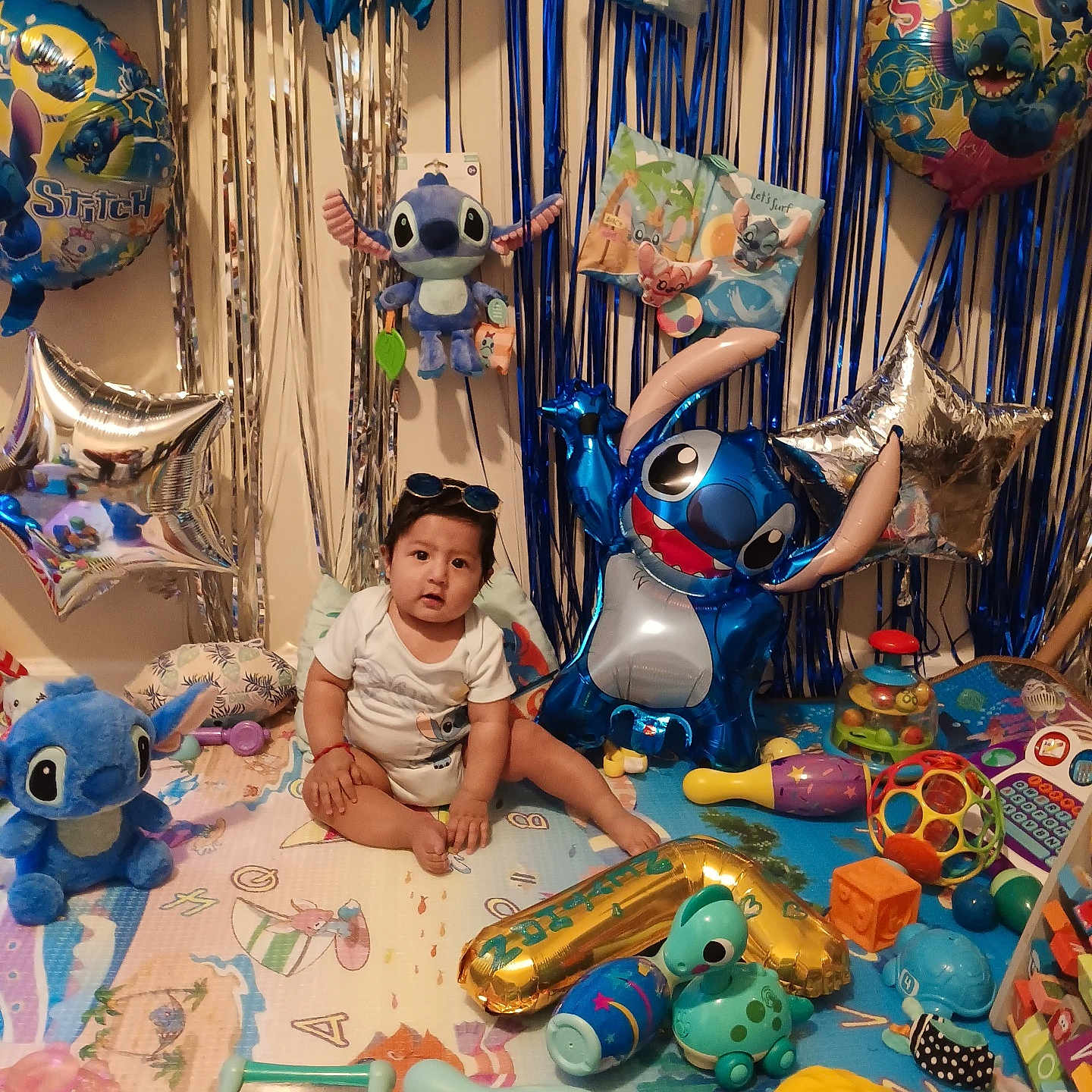 Neythan is registered to the contest to win money with this photo: baby, balloons, birthday, blue, celebration, child, childrens_room, colorful, cute, decorations, floor, indoor, party, plastic_toys, playmat, smile, stitch, stuffed_animals, sunglasses, toys