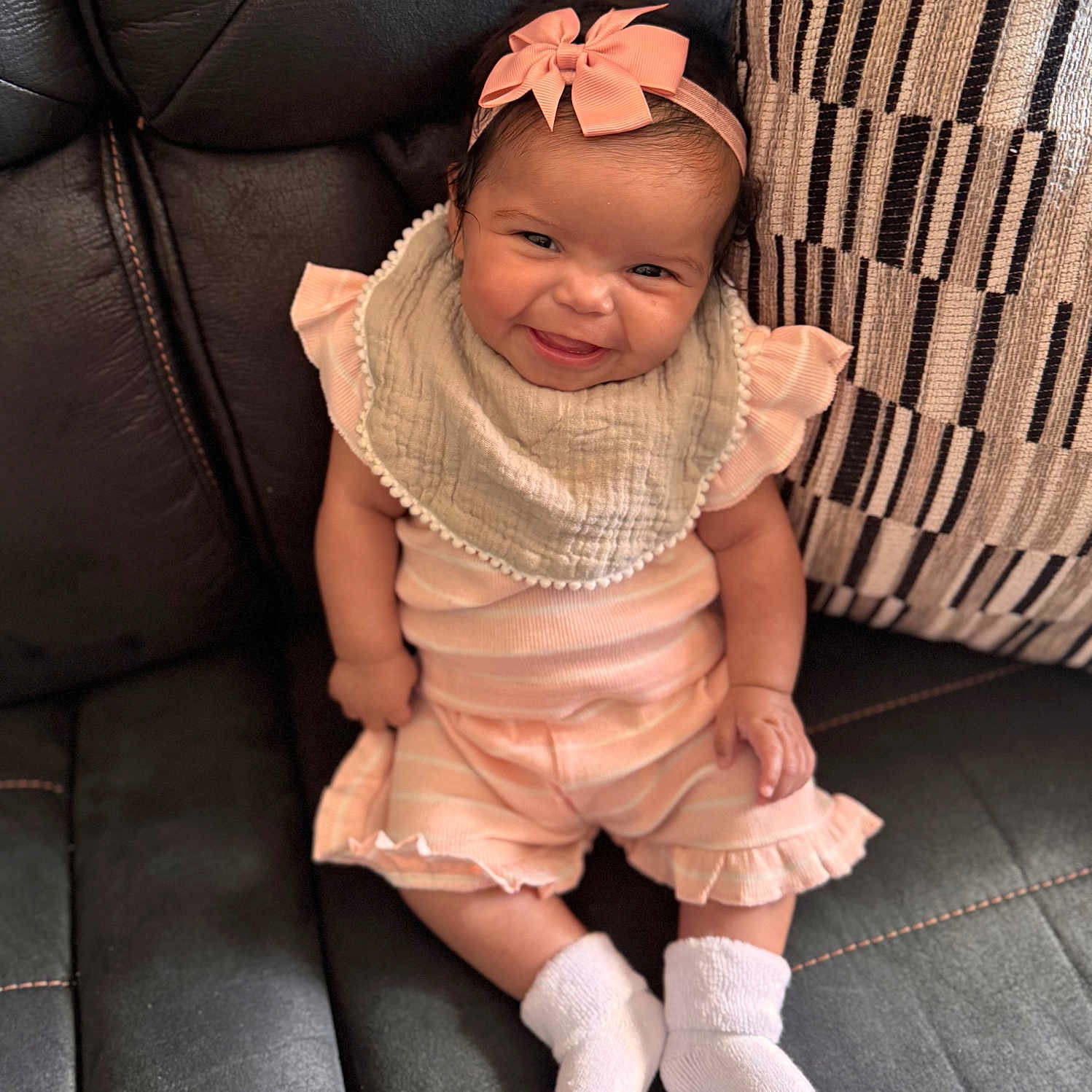 Brynlon is registered to the contest to win money with this photo: baby, bib, black_leather, child, comfortable, couch, cute, happy, headband, indoors, infant, pastel_clothing, patterned_pillow, pillow, pink_bow, portrait, sitting, smiling, sock, young_child