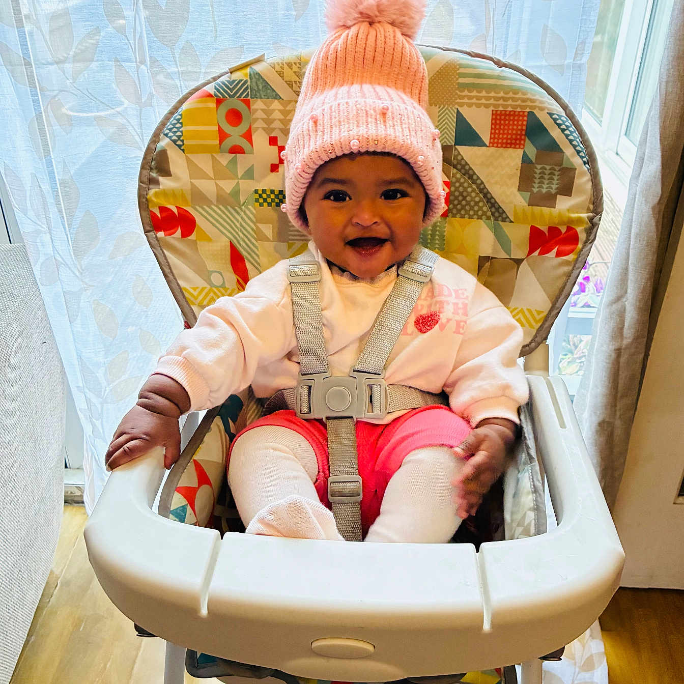 Kiyera is registered to the contest to win money with this photo: baby, child, clothing, colorful, cozy, curtain, cute, floor, happy, high_chair, indoor, person, pink_hat, pom_pom, seat_belt, smiling, toddler, toy, window, wooden_floor