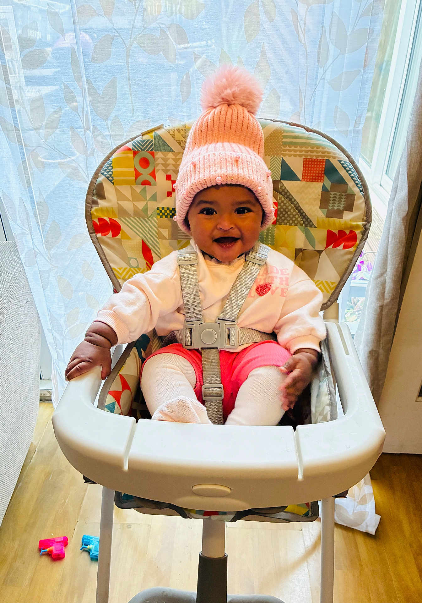 Kiyera is registered to the contest to win money with this photo: baby, high_chair, pink_hat, pom_pom, smiling, happy, indoor, child, toddler, seat_belt, colorful, toy, floor, wooden_floor, curtain, window, clothing, cute, person, cozy