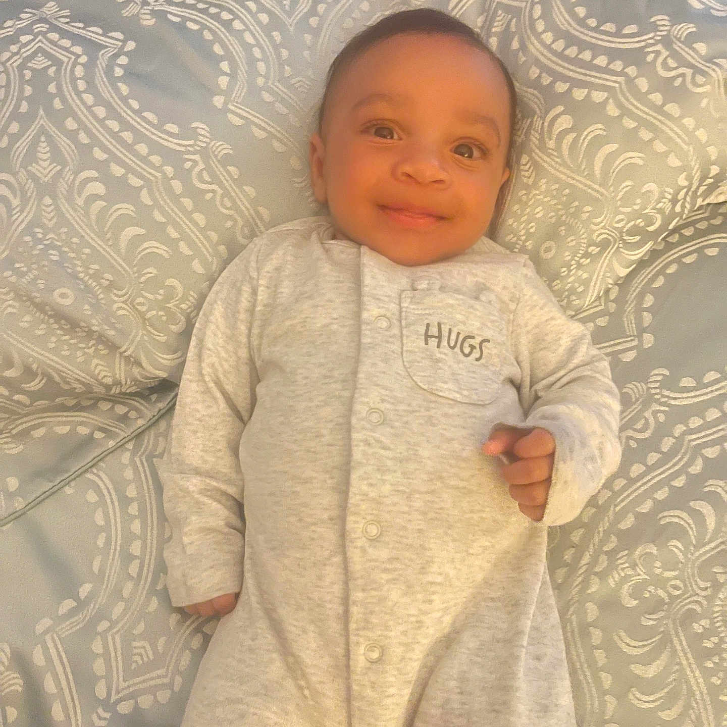 Noah is registered to the contest to win money with this photo: baby, bedspread, child, clothing, comfortable, cute, fabric, face, happy, indoors, infant, lying_down, onesie, pattern, person, portrait, sleepwear, smiling, soft, young