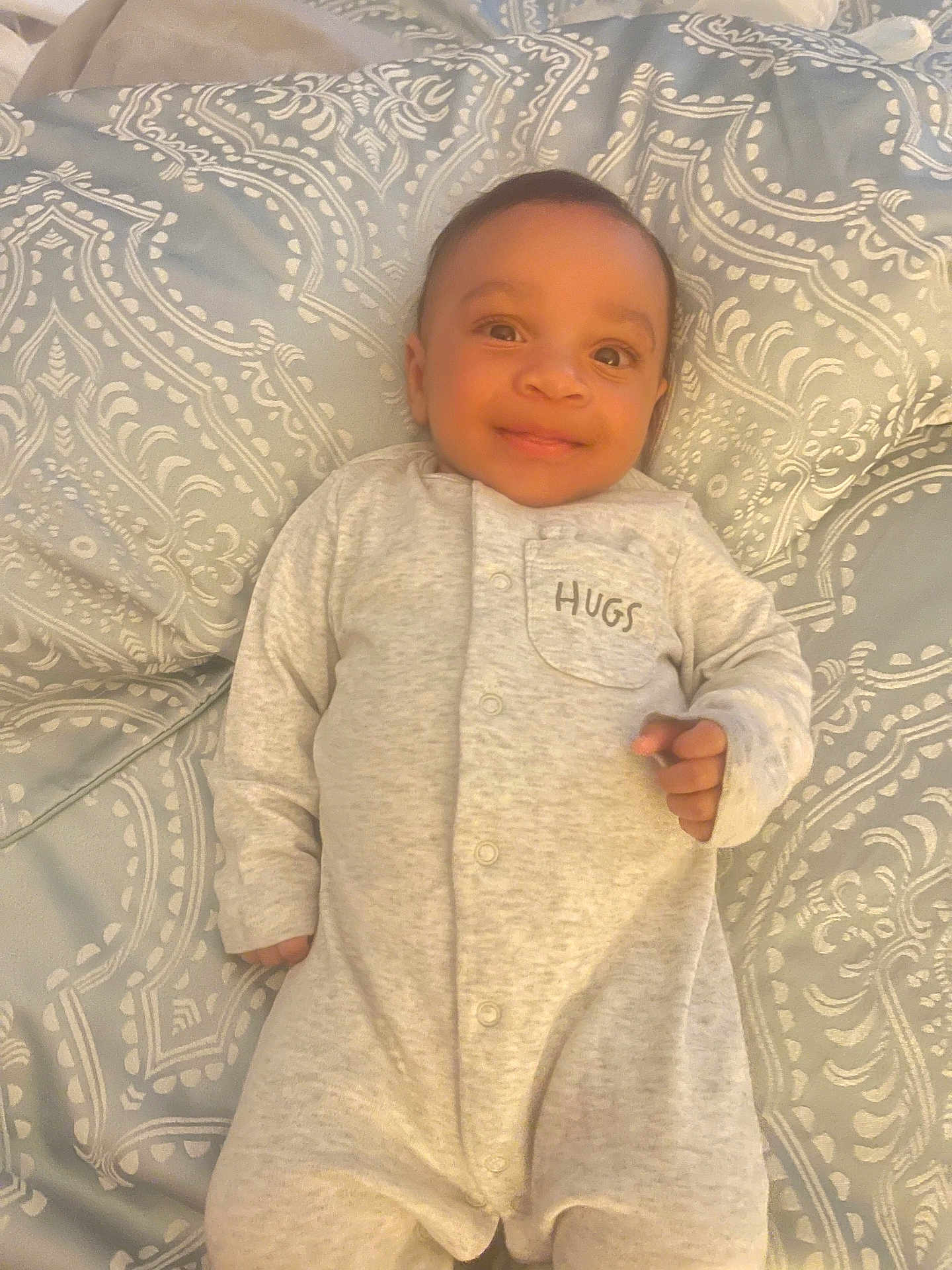 Noah is registered to the contest to win money with this photo: baby, infant, smiling, onesie, clothing, bedspread, pattern, face, person, lying_down, happy, cute, child, young, indoors, fabric, soft, comfortable, portrait, sleepwear