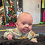 Raven is registered to the contest to win money with this photo: baby, infant, tongue_out, smiling, lying_on_belly, blanket, teddy_print, christmas_tree, holiday_decorations, indoor, mirror_reflection, carpet, cute, portrait, hands, wide_eyes, playful, sweater, floor, person