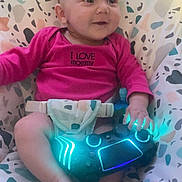 Raven is registered to the contest to win money with this photo: baby, infant, onesie, pink_clothing, bouncer, patterned_fabric, gaming_controller, glowing_lights, hands, feet, face, smile, eyes, seatbelt, indoor, candid, portrait, soft_lighting, cute, child