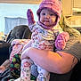 baby, infant, child, pink_hat, mittens, onesie, floral_pattern, holding, adult_arm, couch, living_room, portrait, smile, rosy_cheeks, pajamas, cozy, indoor, seat, blanket, person