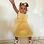 child, curly_hair, cute, fashion, footwear, happy, headband, indoor, person, playful, portrait, raised_hand, sandals, sitting, smiling, stool, tile_floor, toddler, white_wall, yellow_dress