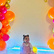 Théa a rejoint le concours — aidez-le/la à gagner de superbes lots ! balloons, birthday_party, child, colorful, cute, decorations, hair_buns, happy, indoor, lighting, long_sleeve_shirt, orange_balloons, pacifier, pink_balloons, pink_dress, sitting, tile_floor, toddler, white_balloons, yellow_balloons