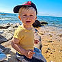 toddler, child, beach, sea, rocks, sand, sunlight, cap, yellow_shirt, shorts, barefoot, outdoor, water, sky, daylight, person, nature, shore, vacation, summer