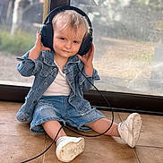 Orsu a rejoint le concours — aidez-le/la à gagner de superbes lots ! blond_hair, cable, casual_clothing, child, curious, cute, daylight, denim_jacket, denim_shorts, floor, glass_door, headphones, indoor, person, sitting, smile, toddler, white_shoes, window, young_child