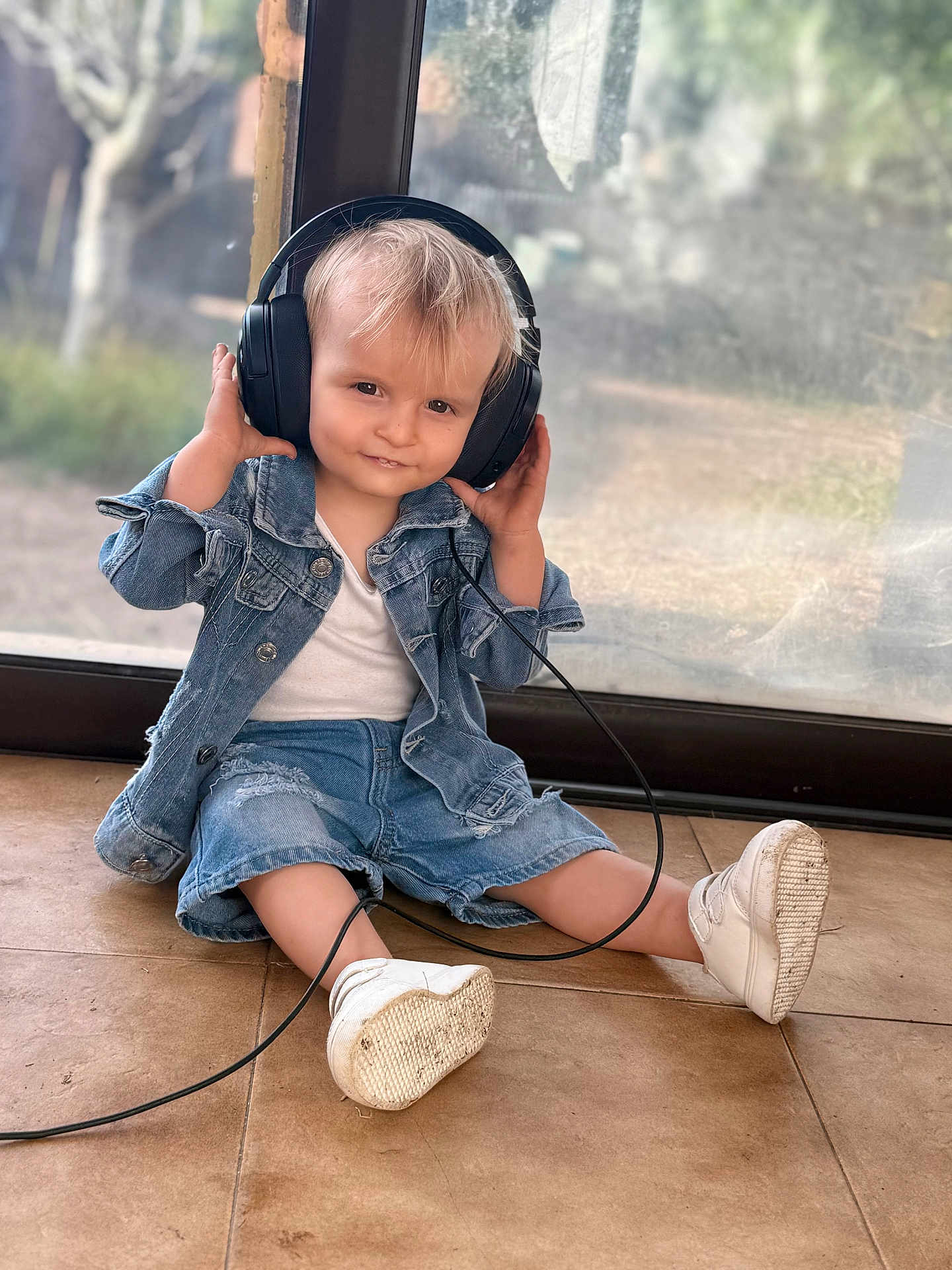 Orsu a rejoint le concours — aidez-le/la à gagner de superbes lots ! blond_hair, cable, casual_clothing, child, curious, cute, daylight, denim_jacket, denim_shorts, floor, glass_door, headphones, indoor, person, sitting, smile, toddler, white_shoes, window, young_child