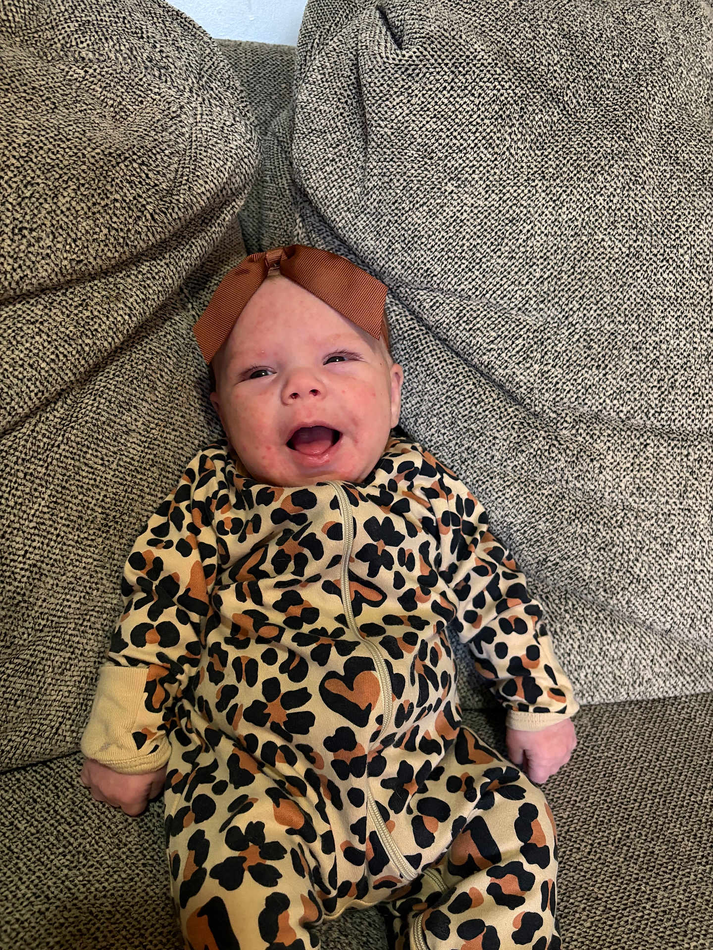 Melody is registered to the contest to win money with this photo: baby, infant, leopard_print, onesie, headband, bow, smiling, happy, sitting, couch, fabric, texture, indoors, child, person, cute, clothing, portrait, young, cozy