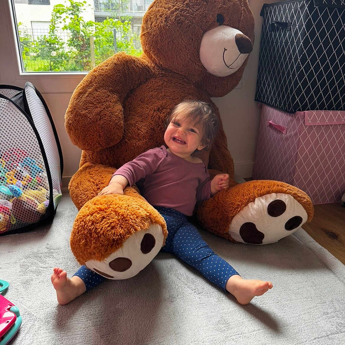 Lou a rejoint le concours — aidez-le/la à gagner de superbes lots ! toddler, child, plush_toy, teddy_bear, carpet, window, indoor, smile, happy, toy_storage, storage_box, play_area, childrens_room, footwear_absent, casual_clothing, natural_light, home, cute, furniture, person