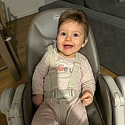 Lou a rejoint le concours — aidez-le/la à gagner de superbes lots ! baby, child, high_chair, smiling, happy, onesie, straps, indoor, living_room, couch, wooden_floor, face, person, seat, clothing, cute, infant, joyful, expression, furniture