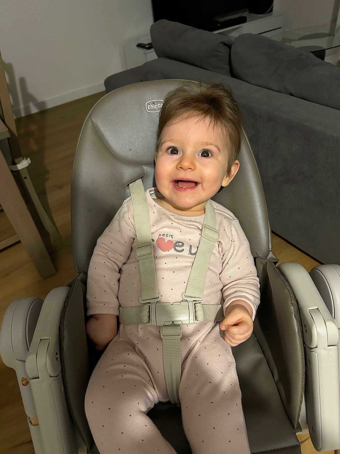 Lou a rejoint le concours — aidez-le/la à gagner de superbes lots ! baby, child, high_chair, smiling, happy, onesie, straps, indoor, living_room, couch, wooden_floor, face, person, seat, clothing, cute, infant, joyful, expression, furniture