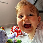 Lou a rejoint le concours — aidez-le/la à gagner de superbes lots ! baby, child, smiling, happy, plush_toy, purple, red_ball, indoor, soft_light, floor, white_clothing, cute, playtime, toy, infant, closeup, face, mouth_open, cheerful, young