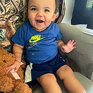 Princtyn is registered to the contest to win money with this photo: toddler, child, smiling, blue_shirt, nike_logo, shorts, teddy_bear, plush_toy, chair, cushion, deer_pillow, indoor, happy, person, legs, hand, face, cute, casual_clothing, home