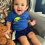 toddler, child, smiling, blue_shirt, nike_logo, shorts, teddy_bear, plush_toy, chair, cushion, deer_pillow, indoor, happy, person, legs, hand, face, cute, casual_clothing, home