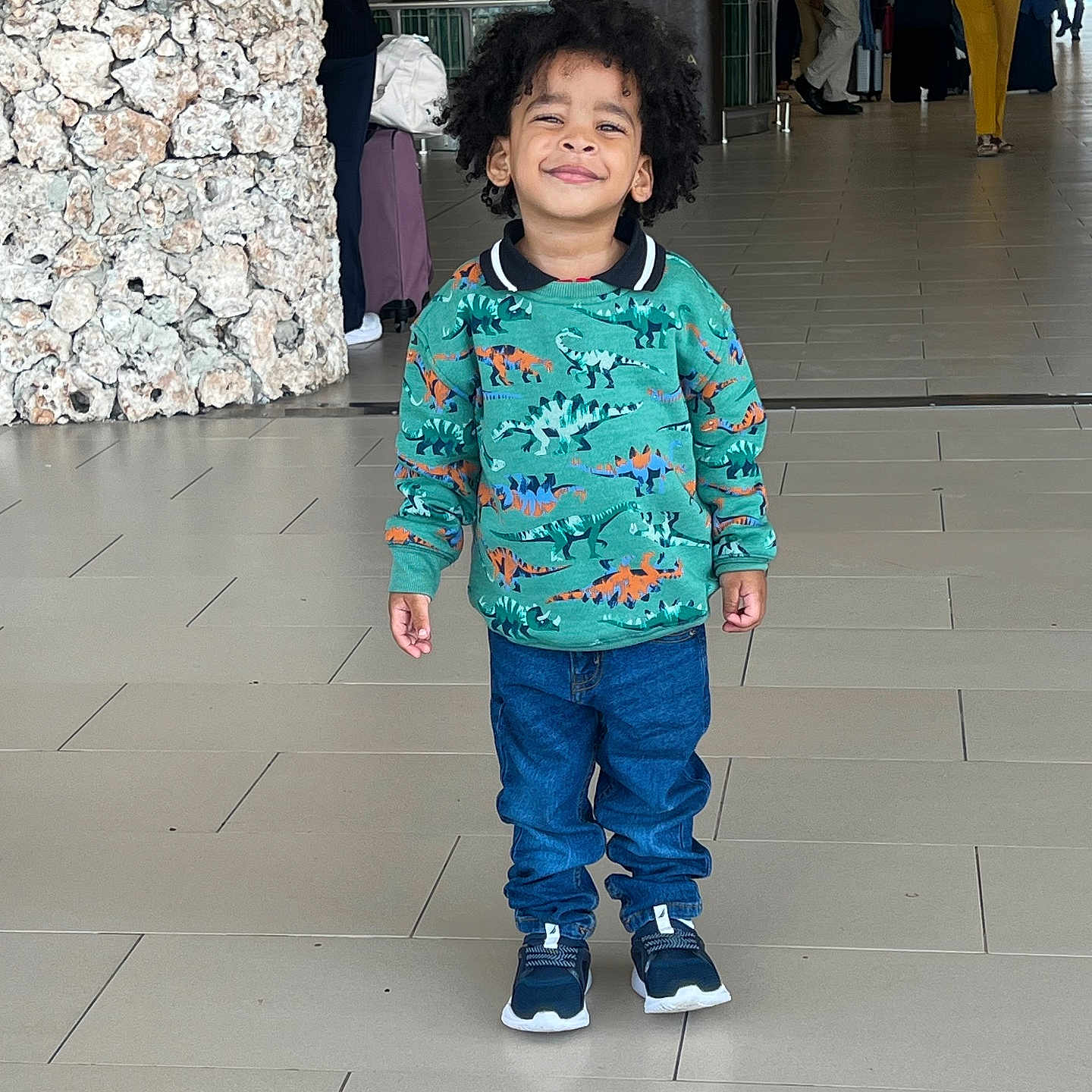 Mateo joined the competition — help win amazing prizes! child, kid, dinosaur_sweater, blue_jeans, sneakers, indoor, public_space, smiling, standing, curly_hair, people, floor_tiles, stone_wall, casual_clothing, portrait, young_child, happy, person, fashion, urban