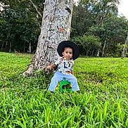 Mateo is registered to the contest to win money with this photo: toddler, child, hat, tree, grass, outdoor, forest, greenery, stool, shirt, dinosaur_print, nature, casual_clothing, young_child, sitting, portrait, daylight, curious_expression, person, scenic