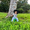 toddler, child, hat, tree, grass, outdoor, forest, greenery, stool, shirt, dinosaur_print, nature, casual_clothing, young_child, sitting, portrait, daylight, curious_expression, person, scenic
