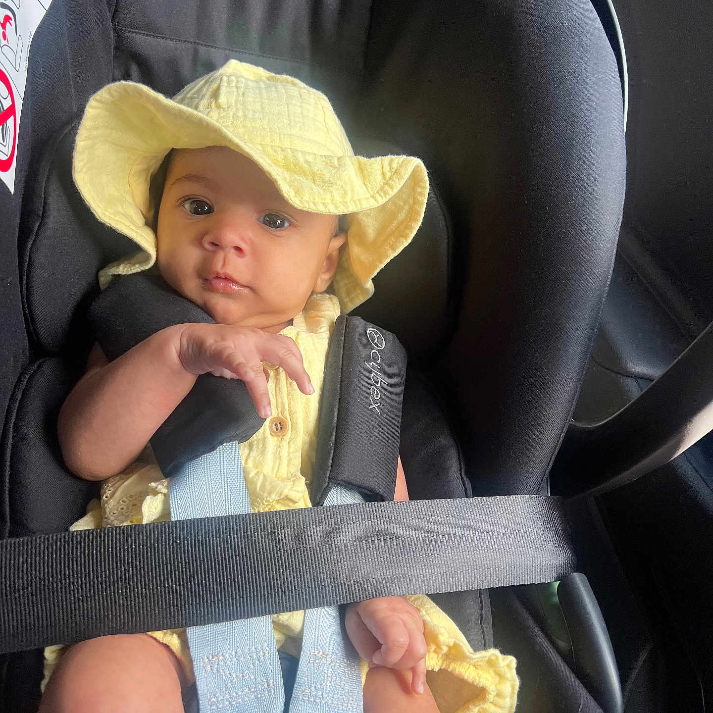 Kiana is registered to the contest to win money with this photo: baby, black_seat, car_interior, car_seat, car_trip, child, child_safety, cute, hat, infant, infant_clothing, looking_at_camera, portrait, safety, seatbelt, small_hands, soft_light, straps, yellow_clothing, yellow_hat