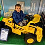 toddler, child, toy_car, yellow, blue_jacket, indoor, store_display, steering_wheel, curious, vehicle, plastic, small_wheels, play_area, person, shopping, floor, green_carpet, face, clothing, pose