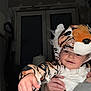toddler, child, costume, tiger, animal_print, cute, smiling, hand, indoor, window, adult_hand, dark_background, playful, face, headwear, clothing, person, portrait, baby, fun