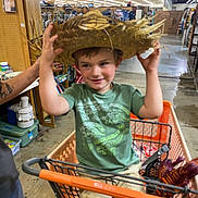 Dawson is registered to the contest to win money with this photo: child, boy, shopping_cart, hat, straw_hat, green_shirt, dinosaur_print, shorts, indoor, store, warehouse, toys, smiling, person, hand, floor, shelf, boxes, lighting, playful
