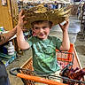 child, boy, shopping_cart, hat, straw_hat, green_shirt, dinosaur_print, shorts, indoor, store, warehouse, toys, smiling, person, hand, floor, shelf, boxes, lighting, playful