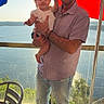 child, toddler, man, adult, holding, umbrella, outdoor, lake, water, sunlight, smile, casual_clothing, jeans, white_shoes, railing, chair, nature, portrait, family, happy